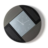 Orologio da parete Oblique dal design geometrico e originale, con struttura circolare in ferro e caratteristico quadrante a rombo in vetro specchiato. Vista frontale, finitura fango/nero/fumé.